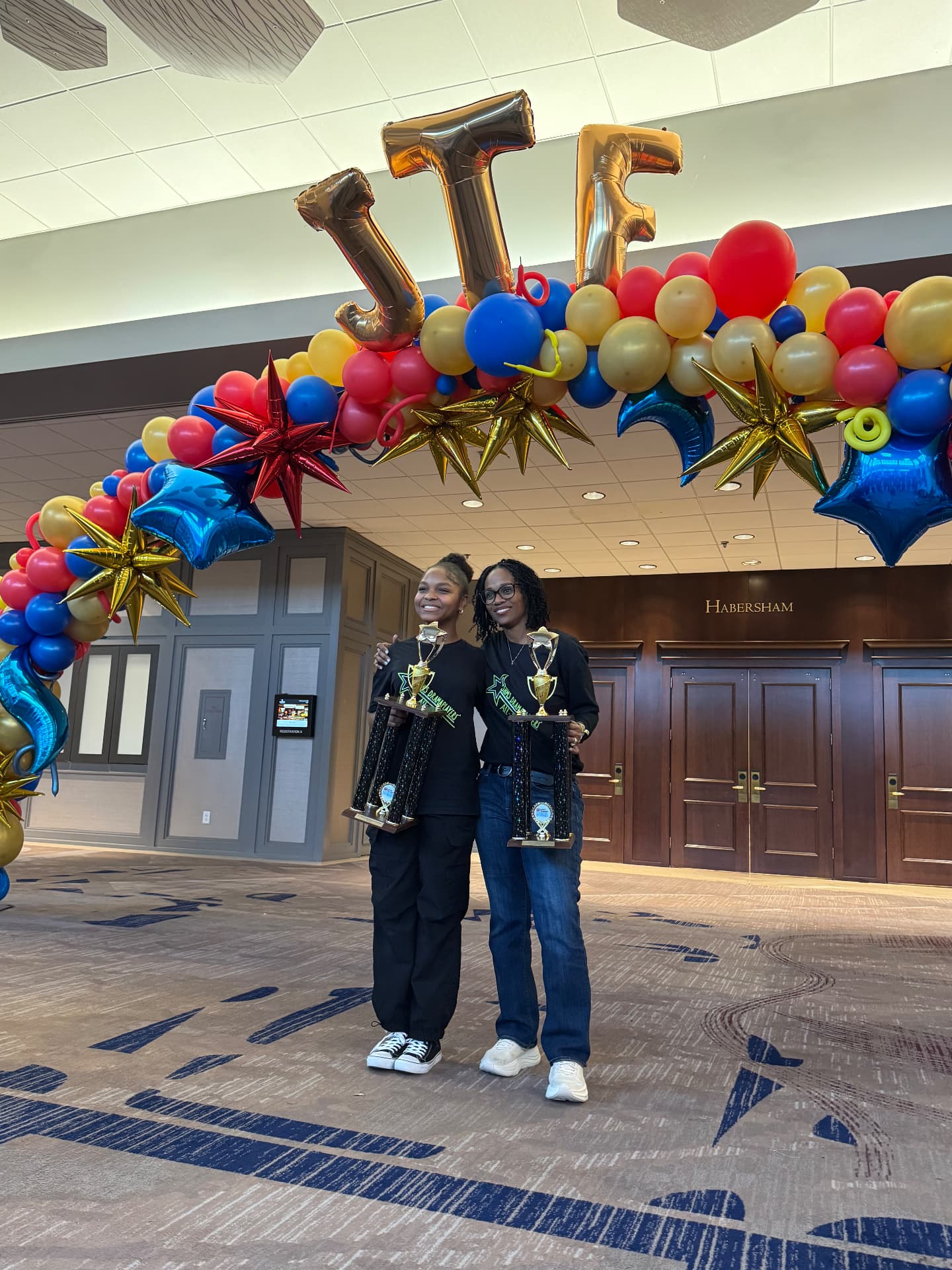 Asherah and her director holding trophies beneath a festival balloon arch